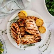 Grilled lemon chicken breasts on a white plate with charred lemon halves and sprigs of thyme, surrounded by a cloth napkin, greens, and a small dish of asparagus.