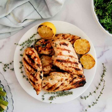 Grilled lemon chicken breasts on a white plate with charred lemon halves and sprigs of thyme, surrounded by a cloth napkin, greens, and a small dish of asparagus.