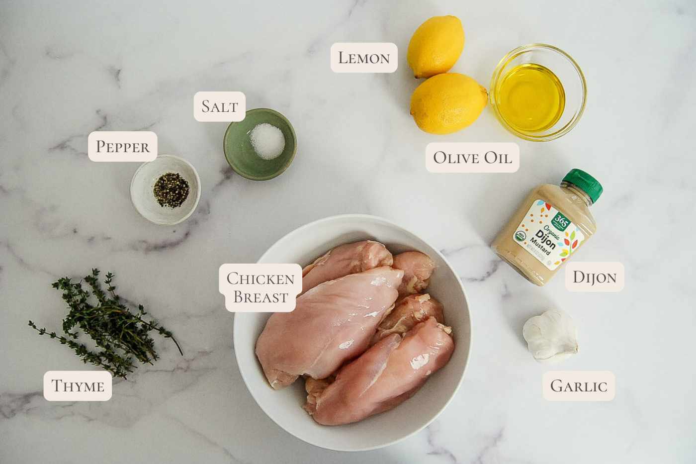 Raw chicken breasts in a bowl, with labeled ingredients around: thyme, pepper, salt, lemon, olive oil, Dijon mustard, and garlic, all on a white marble surface.