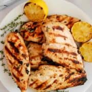 Grilled lemon chicken breasts with char marks, garnished with fresh thyme and grilled lemon halves, served on a white plate with a light cloth in the background.