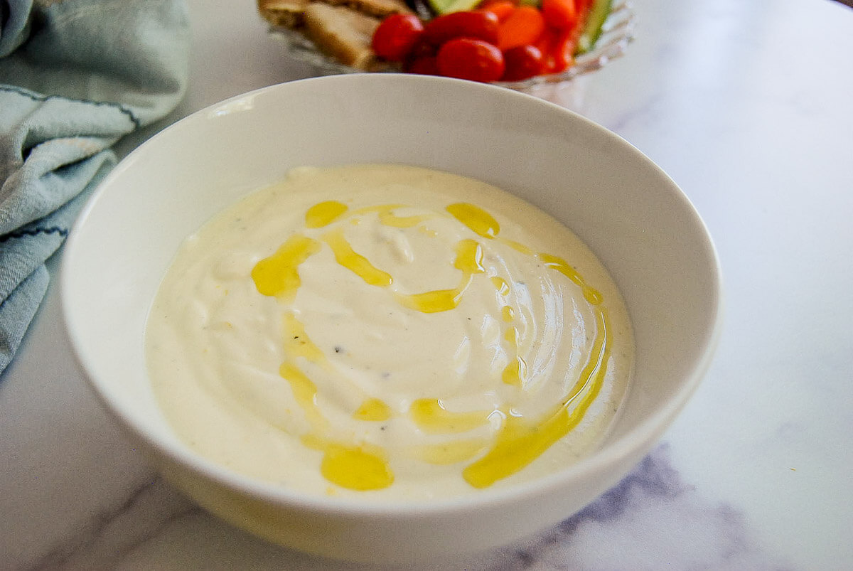 A white bowl filled with whipped feta drizzled with olive oil, sitting on a marble surface, with a plate of vegetables and bread blurred in the background.
