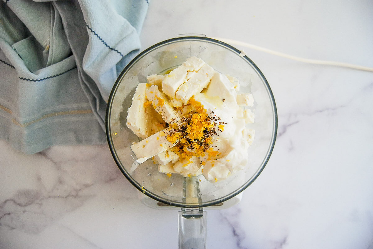 Overhead view of a food processor containing blocks of feta cheese, yogurt, lemon zest, and black pepper.