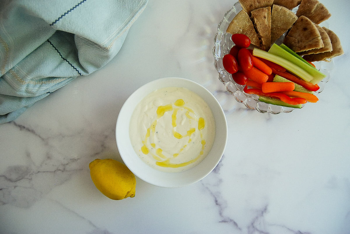 A bowl of creamy whipped feta dip drizzled with olive oil, next to a lemon, pita bread slices, cherry tomatoes, celery, and carrot sticks.