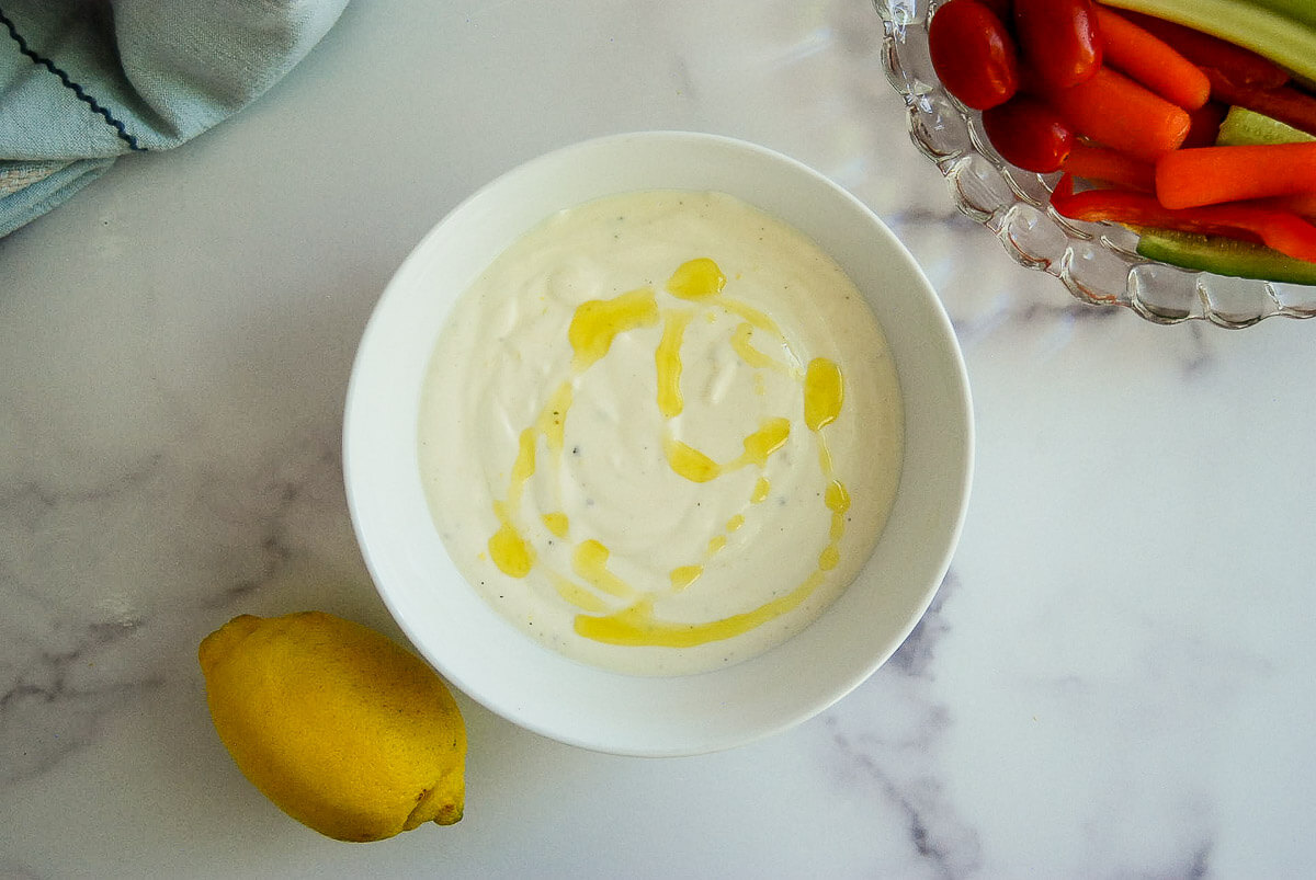 A bowl of creamy whipped feta dip drizzled with olive oil sits on a marble surface next to a lemon and a platter of fresh vegetables including carrots, celery, and grape tomatoes.