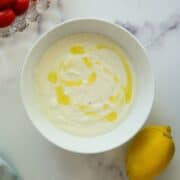 A bowl of whipped feta with olive oil drizzled on top, next to a lemon and a partial view of cherry tomatoes on a white marble surface.