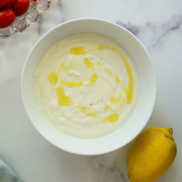A bowl of whipped feta with olive oil drizzled on top, next to a lemon and a partial view of cherry tomatoes on a white marble surface.