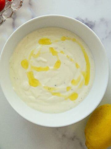 A bowl of whipped feta with olive oil drizzled on top, next to a lemon and a partial view of cherry tomatoes on a white marble surface.