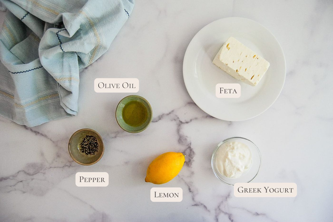 Marble counter with labeled ingredients: olive oil, feta on a plate, a bowl of Greek yogurt, a whole lemon, ground pepper in a bowl, and a blue kitchen towel.