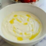A white bowl of creamy whipped feta dip drizzled with olive oil, with a platter of sliced bread, celery, cherry tomatoes, and carrot sticks blurred in the background.