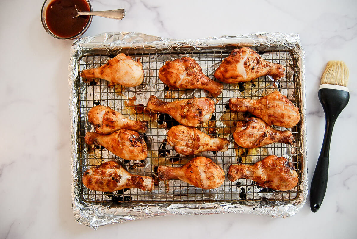 Twelve baked bbq chicken drumsticks on a wire rack over a foil-lined baking sheet, with a small bowl of barbecue sauce and a basting brush next to them.