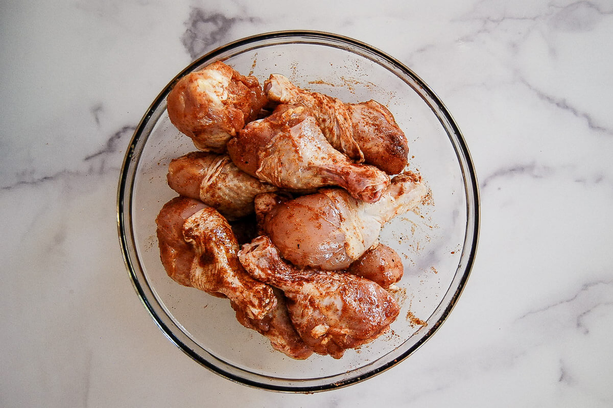 Raw chicken drumsticks coated in spices are arranged in a glass bowl on a white marble surface.