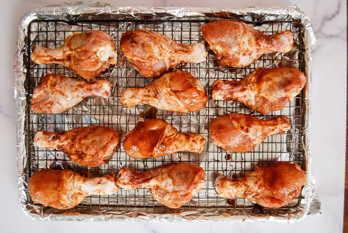Twelve raw, seasoned chicken drumsticks are arranged on a wire rack over a foil-lined baking sheet, ready for cooking.