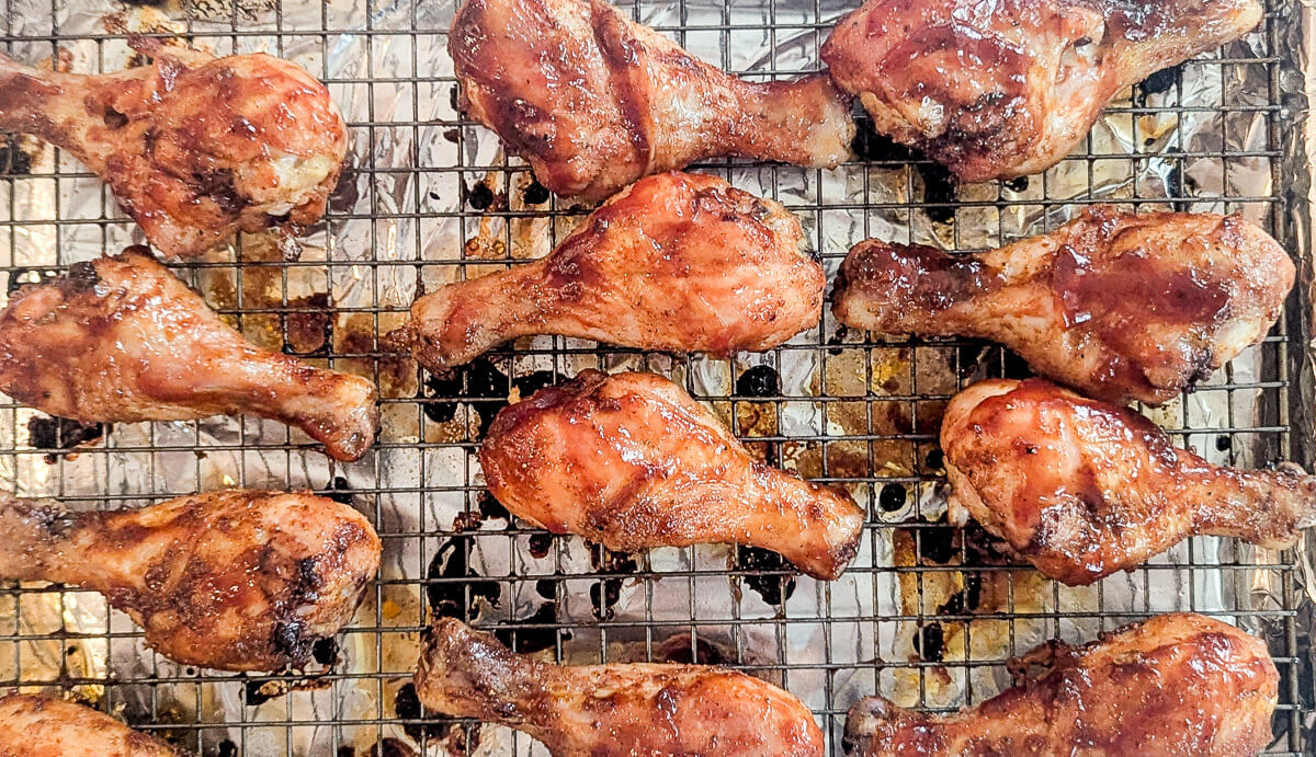 Twelve glazed bbq chicken drumsticks arranged on a metal wire rack over a baking sheet, with some char marks and caramelized sauce visible on the skin.