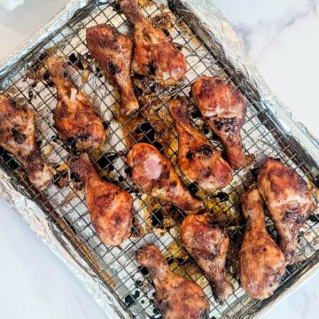 Twelve baked bbq chicken drumsticks on a wire rack over a foil-lined baking sheet, with visible seasoning and browned skin.
