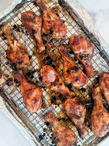 Twelve baked bbq chicken drumsticks on a wire rack over a foil-lined baking sheet, with visible seasoning and browned skin.
