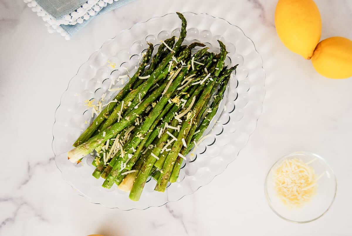 Roasted asparagus spears with lemon on a clear glass plate, garnished with grated cheese. Two whole lemons and a small bowl of grated cheese sit nearby on a white marble surface.