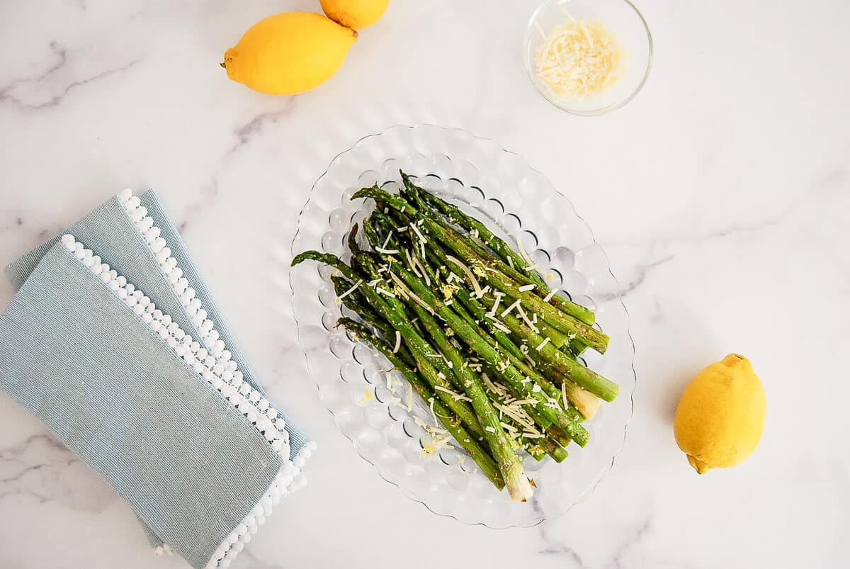 A glass platter of roasted asparagus topped with shredded cheese sits on a marble surface. Nearby are two lemons, a blue napkin, and a small bowl of grated cheese.