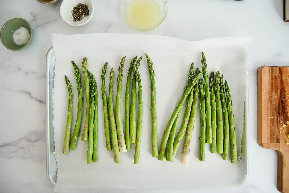 Fresh asparagus spears arranged in a single layer on a parchment-lined baking sheet, ready to be cooked. Nearby are bowls of seasoning, lemon juice, and a wooden board with minced garlic.