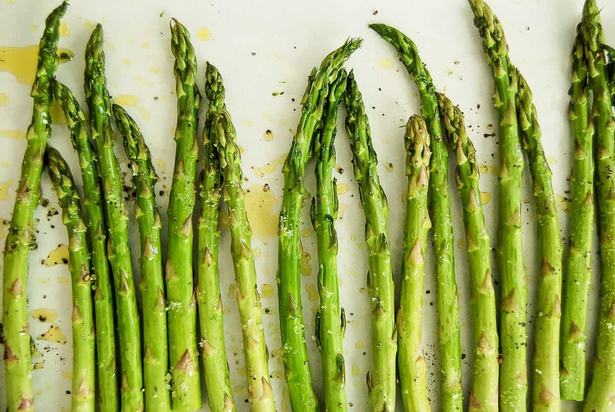 Fresh asparagus spears arranged in rows on a white surface, drizzled with olive oil and sprinkled with black pepper and herbs.
