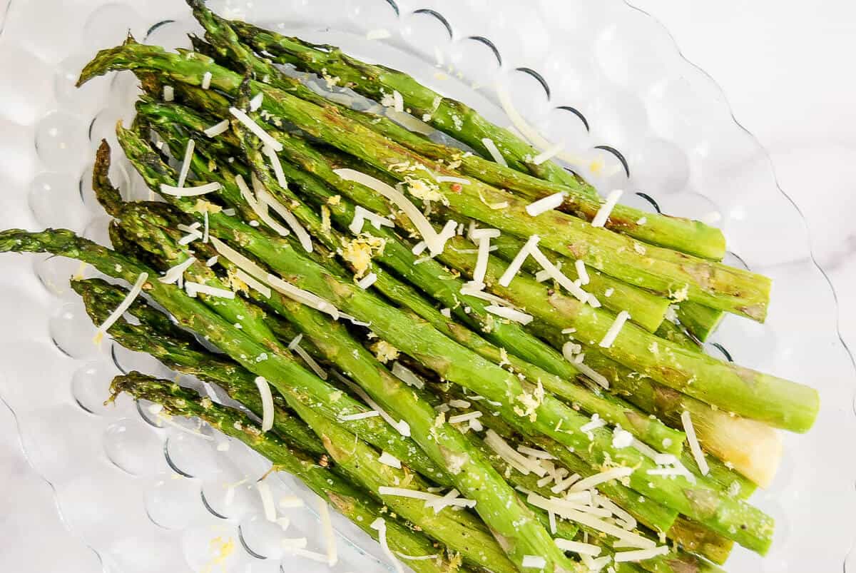 Roasted asparagus spears with lemon topped with shredded parmesan cheese, served on a clear, textured glass plate.