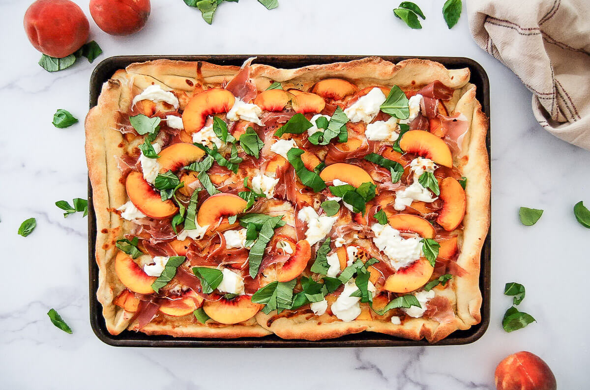 Rectangular pizza topped with peach slices, fresh basil, prosciutto, and dollops of cheese on a baking sheet.