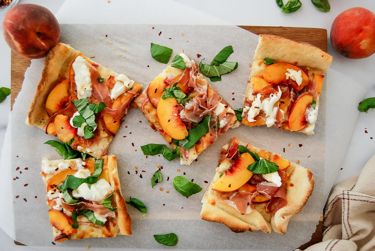 Five rectangular peach, prosciutto, and burrata pizza slices are arranged on parchment paper, garnished with fresh basil. Whole peaches and scattered basil leaves surround the slices.