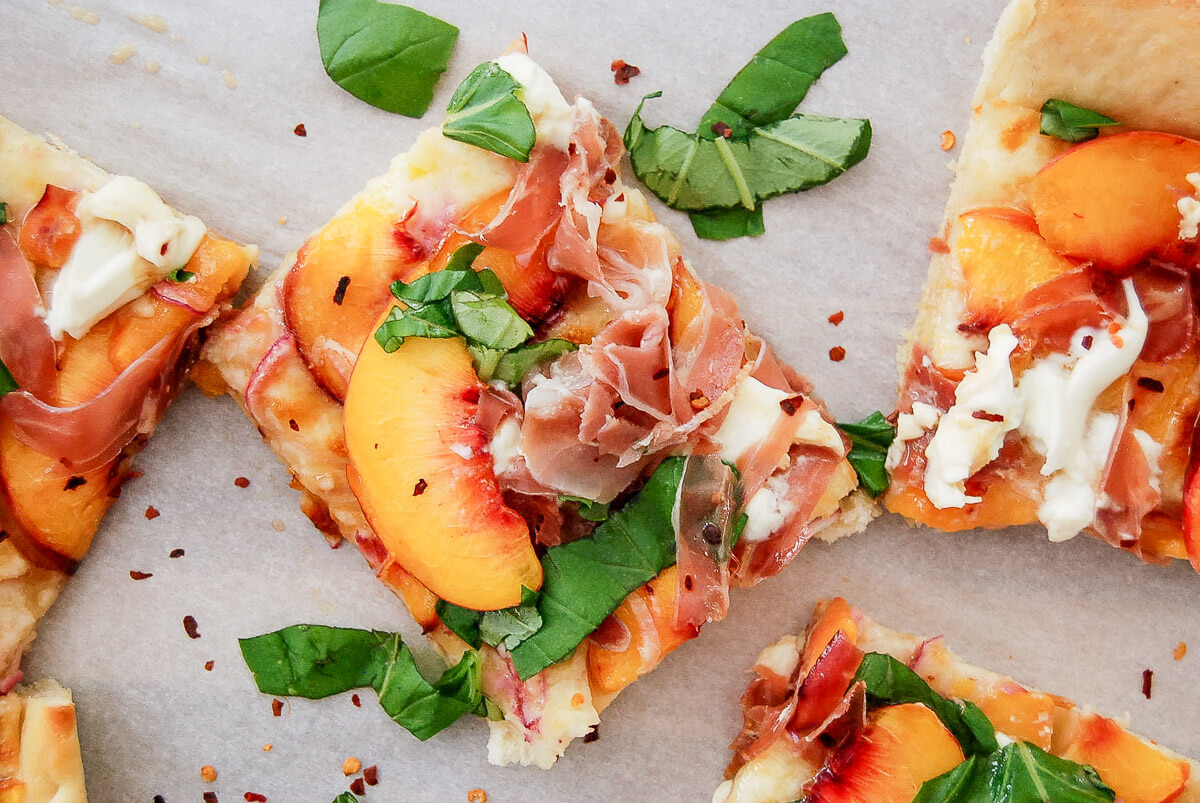 Peach pizza slices topped with peach slices, prosciutto, fresh basil, cheese, and red pepper flakes on parchment paper.