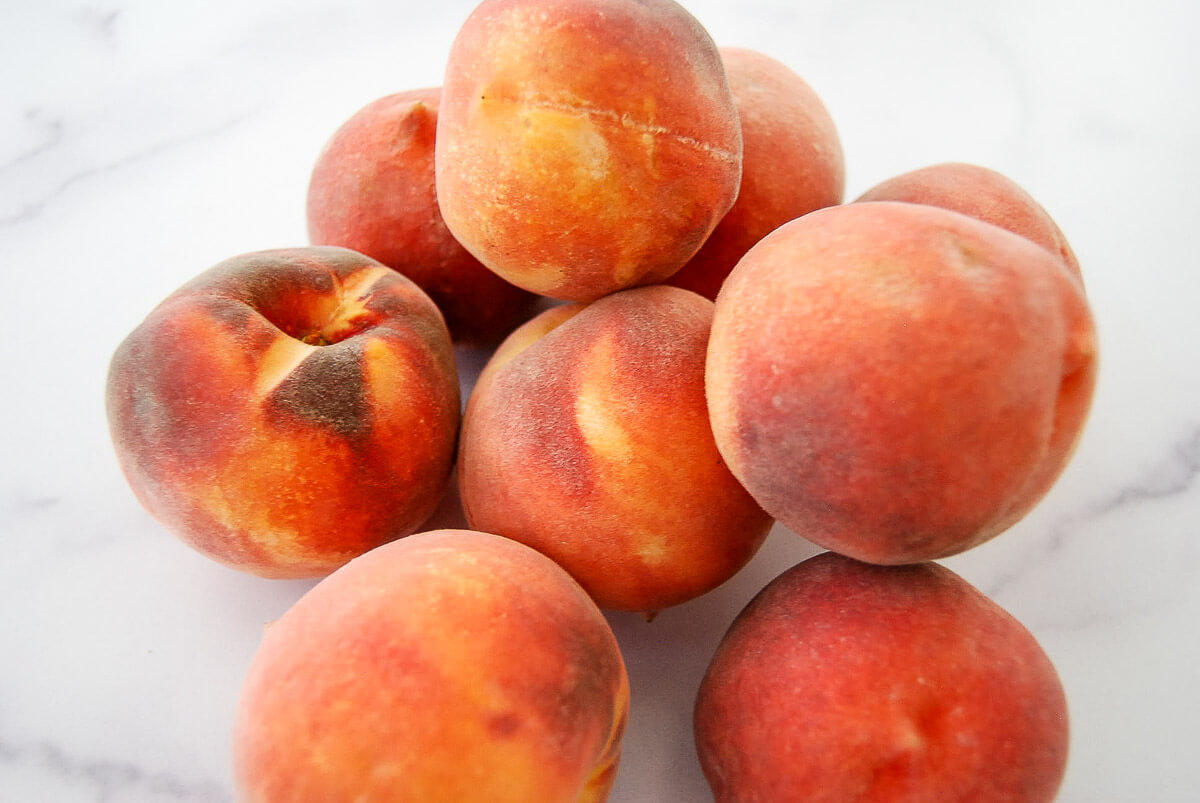 A group of ripe peaches with reddish-orange skin rests on a white surface.
