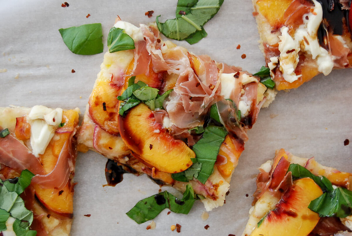 Rectangular slices of peach pizza topped with prosciutto, fresh peach slices, basil leaves, mozzarella, and a drizzle of balsamic glaze on parchment paper.
