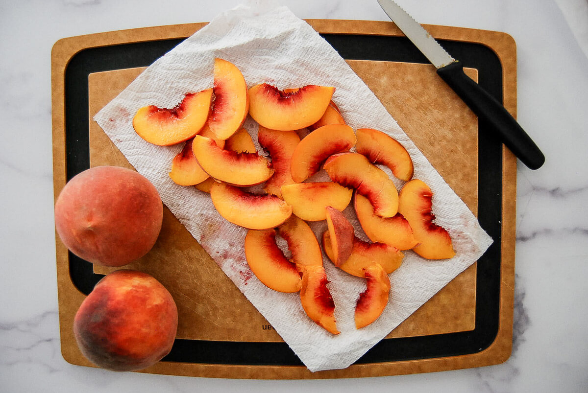 Two whole peaches and sliced peaches on a paper towel rest on a cutting board, with a knife placed nearby on a marble surface.