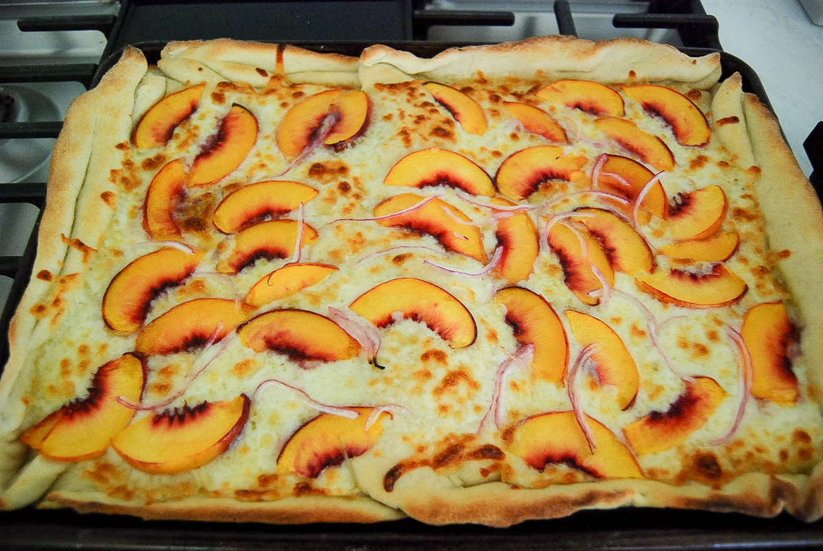 Rectangular pizza topped with melted cheese, thin peach slices, and slivers of red onion on a baking tray.