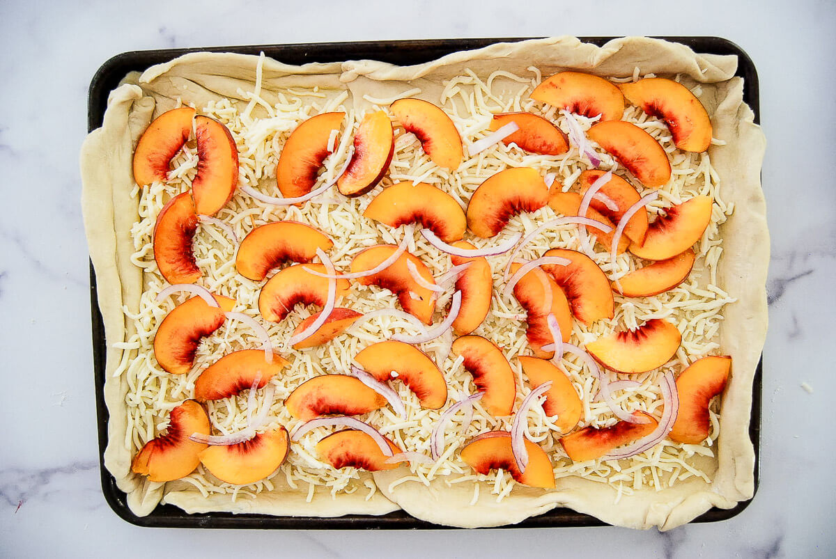 Rectangular pizza dough in a pan topped with shredded cheese, peach slices, and thin red onion slices.