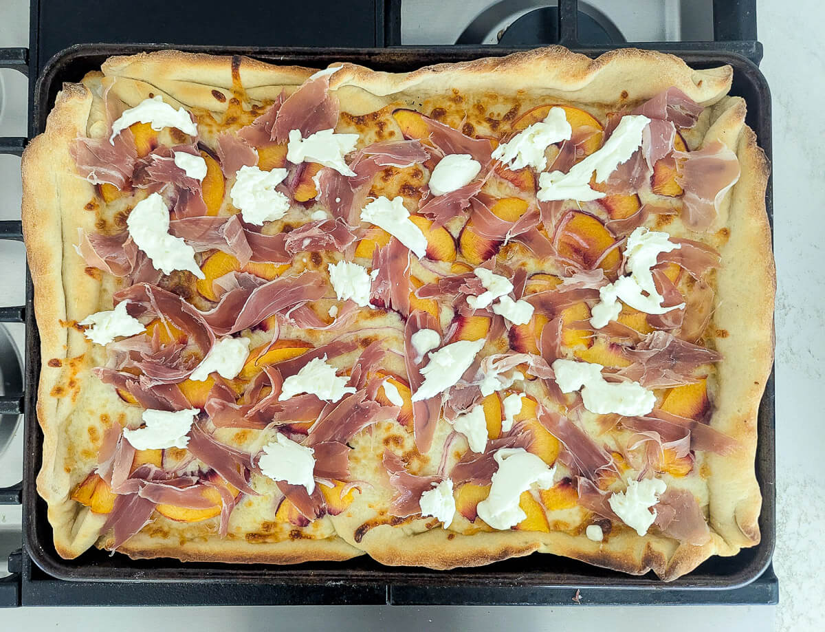 Rectangular pizza topped with peach slices, prosciutto, and dollops of burrata on a baking tray.
