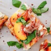 A square slice of peach pizza topped with prosciutto, peach slices, fresh basil leaves, and melted cheese, garnished with red pepper flakes.