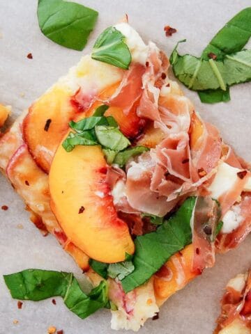 A square slice of peach pizza topped with prosciutto, peach slices, fresh basil leaves, and melted cheese, garnished with red pepper flakes.