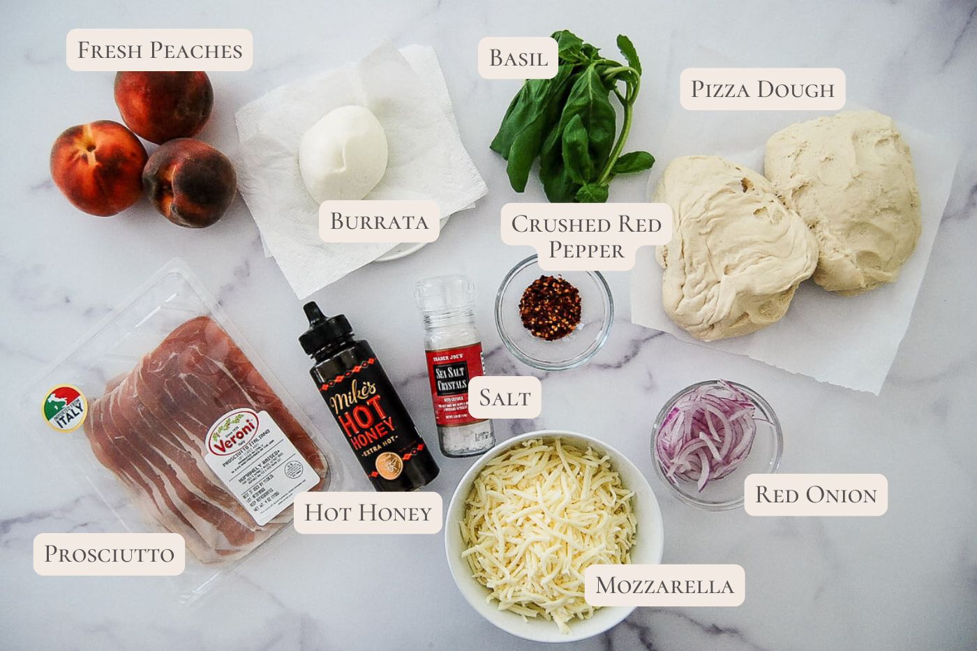 Peach pizza ingredients laid out on a white surface: fresh peaches, burrata, basil, pizza dough, crushed red pepper, salt, hot honey, prosciutto, mozzarella, and sliced red onion, each labeled with text.