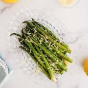 Roasted asparagus spears topped with grated cheese and lemon zest, arranged on a clear glass plate on a white marble surface.
