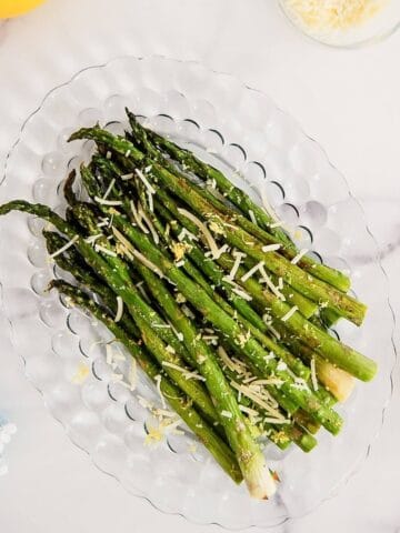 Roasted asparagus spears topped with grated cheese and lemon zest, arranged on a clear glass plate on a white marble surface.