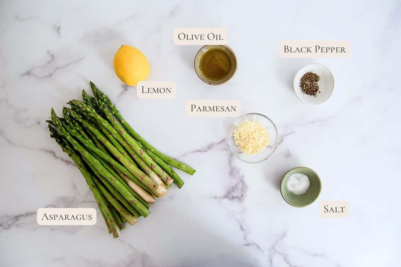Several ingredients labeled on a marble surface: a bunch of asparagus, a lemon, bowls of olive oil, grated parmesan, black pepper, and salt.