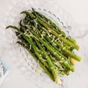 Roasted asparagus spears are arranged on a clear glass plate, topped with shredded parmesan cheese and lemon zest. The plate sits on a white marble surface.