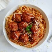 A white bowl of spaghetti topped with tomato sauce, turkey meatballs, grated cheese, and a basil garnish, on a white surface with a fork and a napkin nearby.
