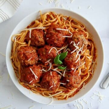A white bowl of spaghetti topped with tomato sauce, turkey meatballs, grated cheese, and a basil garnish, on a white surface with a fork and a napkin nearby.