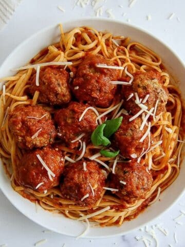 A white bowl of spaghetti topped with tomato sauce, turkey meatballs, grated cheese, and a basil garnish, on a white surface with a fork and a napkin nearby.