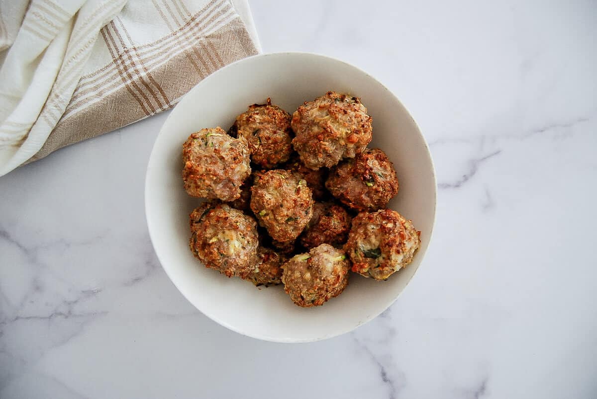 A white bowl filled with homemade baked turkey meatballs sits on a marble surface next to a folded beige and white kitchen towel.