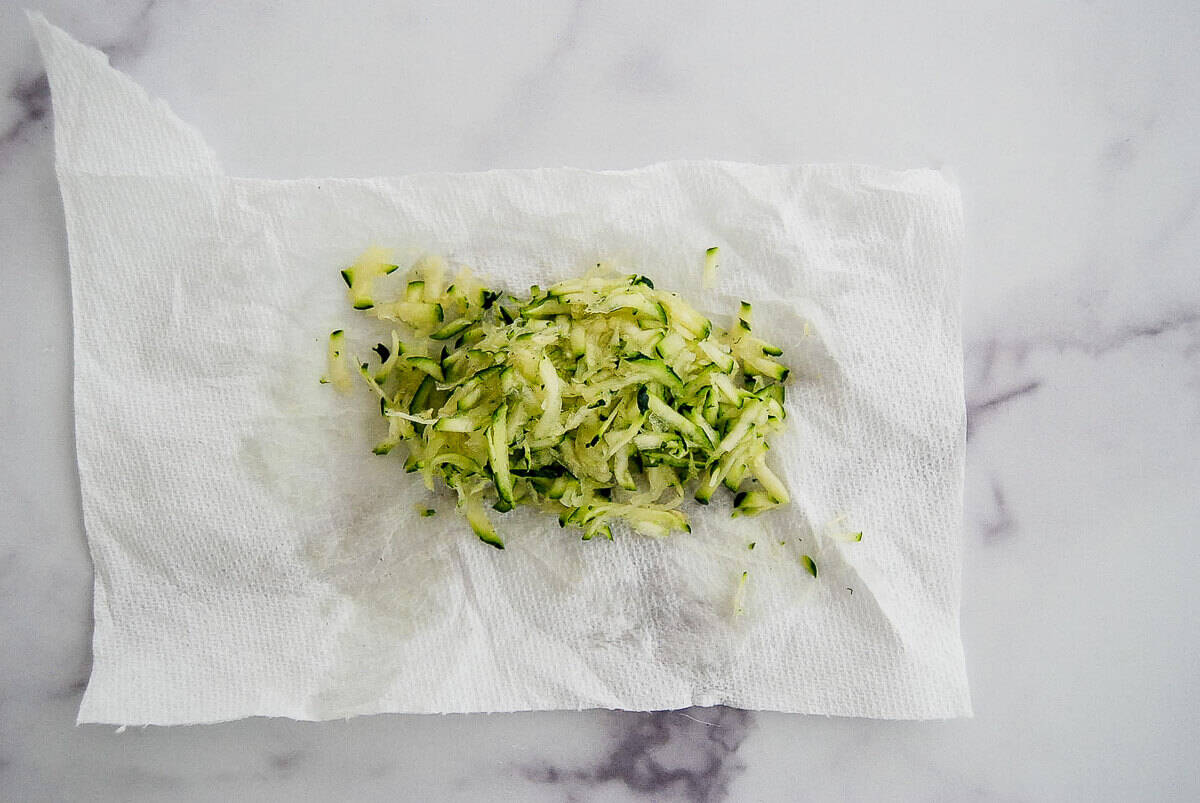 Shredded zucchini sits on a white paper towel on a marble surface, with moisture visible on the towel.