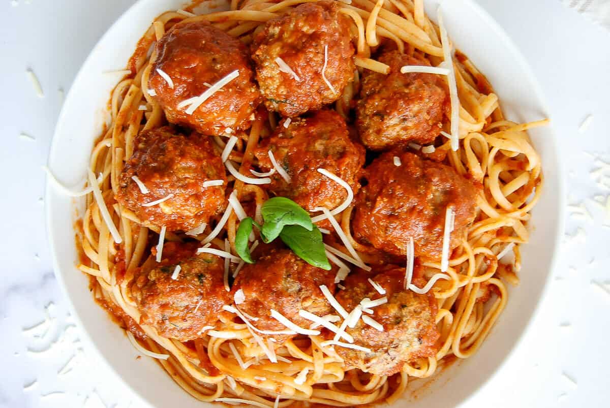 A white plate filled with spaghetti topped with baked turkey meatballs in tomato sauce, garnished with shredded cheese and a sprig of fresh basil in the center.