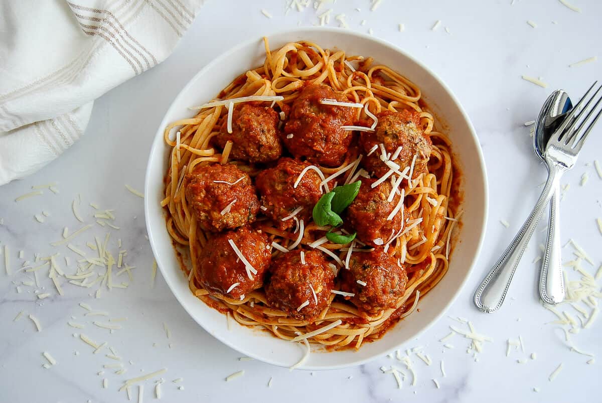 A bowl of spaghetti with tomato sauce, turkey meatballs, grated cheese, and a basil garnish. A fork and spoon are beside the bowl on a white surface sprinkled with cheese.