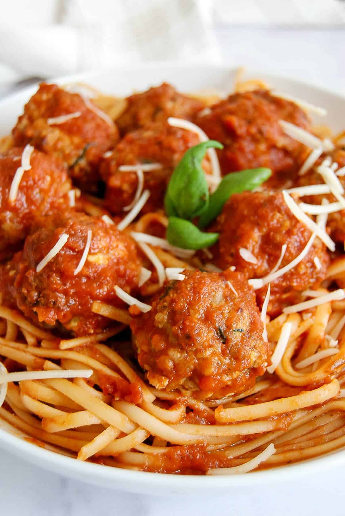 A plate of spaghetti topped with baked turkey meatballs, marinara sauce, shredded cheese, and a sprig of fresh basil.