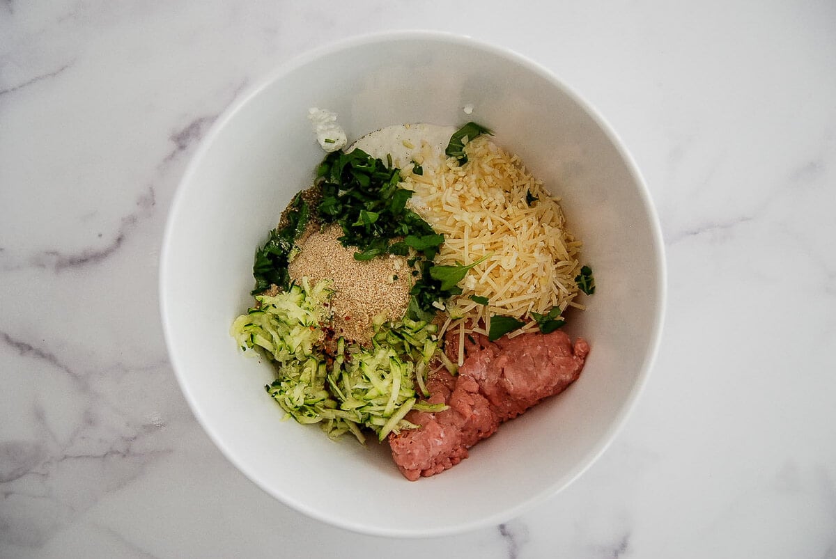 White bowl containing ground turkey, shredded zucchini, grated cheese, breadcrumbs, chopped herbs, and cottage cheese, all separated in sections on a white marble surface.
