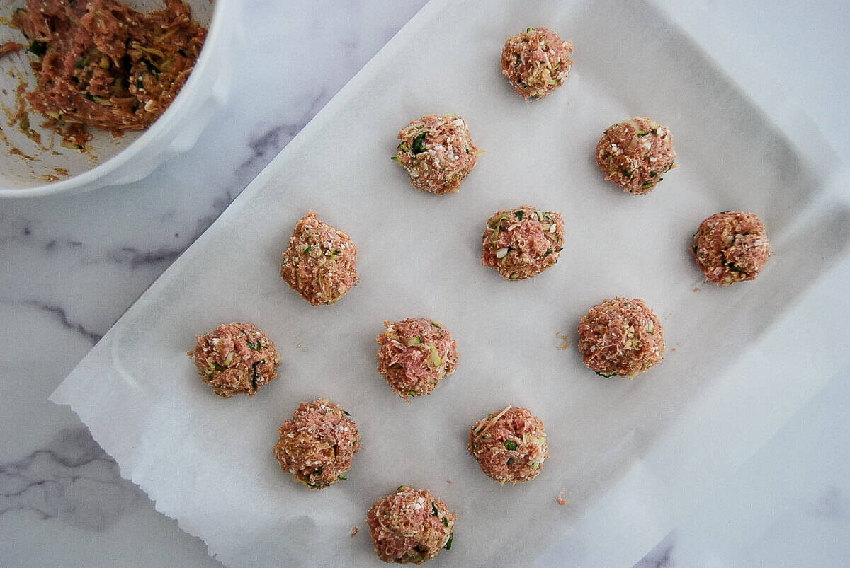 Raw turkey meatballs arranged on a parchment-lined baking sheet, with a bowl of meat mixture in the upper left corner on a white marble surface.